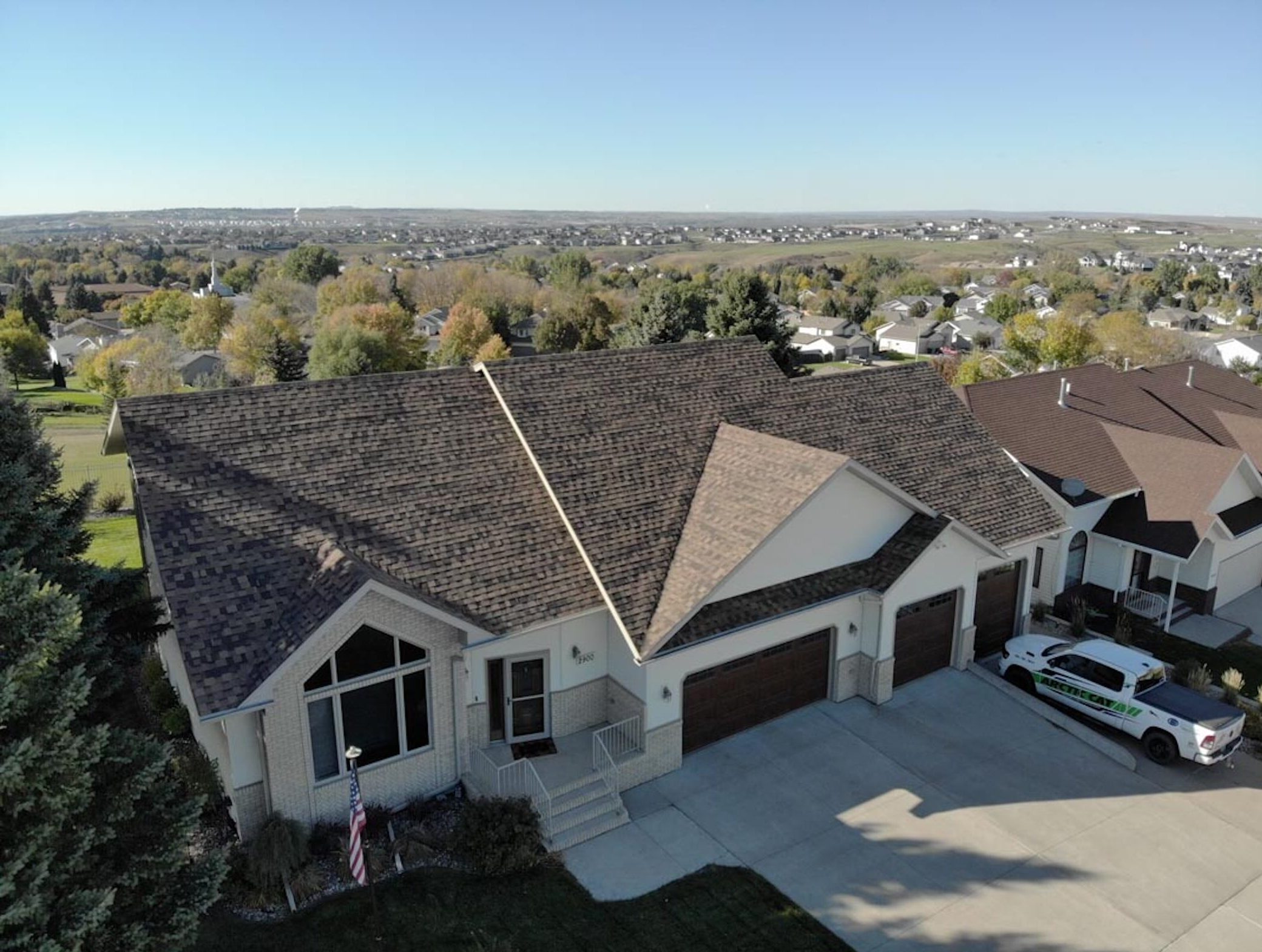 Aerial view of a suburban home with a newly installed shingle roof, surrounded by trees with fall foliage and neighboring houses, under a clear blue sky.